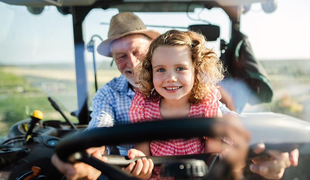 A senior farmer with small granddaughter sitting on his lap while they drive a tractor, preparing for the next CSA pickup.