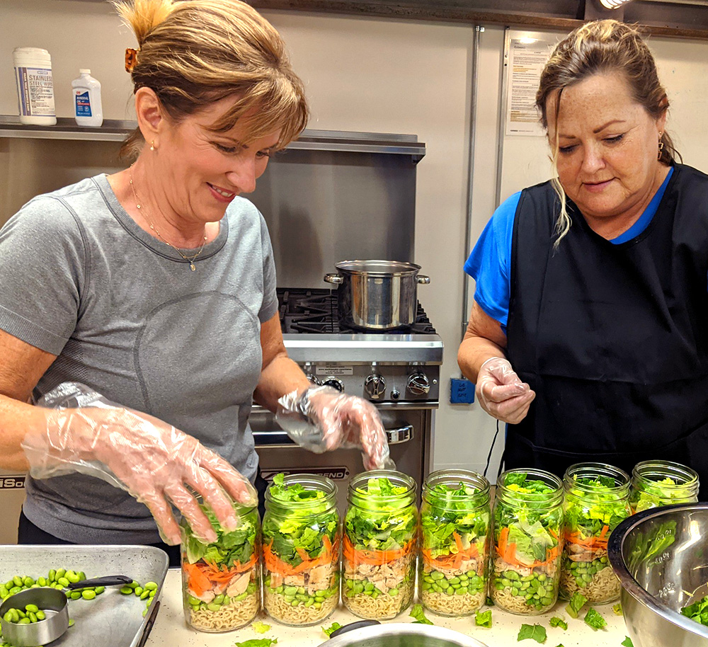 Two women volunteers fill glass jars with fresh greens as an option for prepared foods at prairieland market